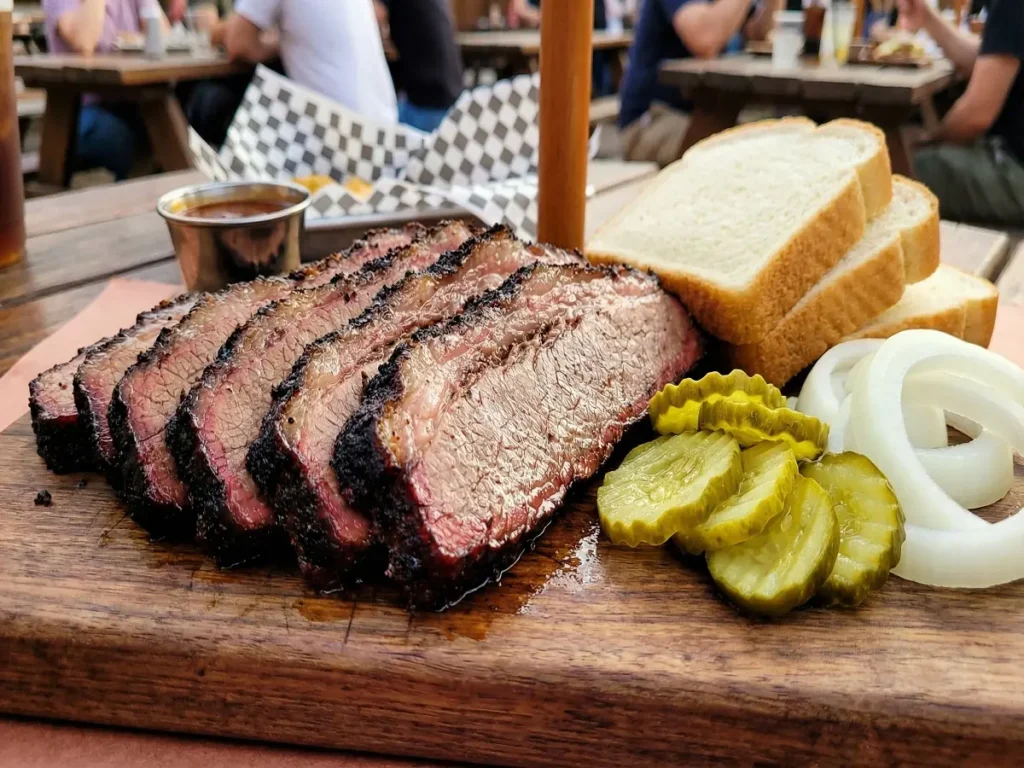 Sliced smoked brisket against the grain served with pickles onions and white bread
